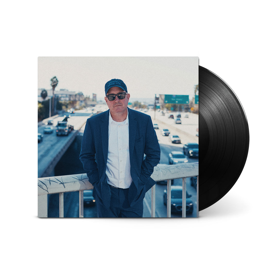 A Craig Finn vinyl record features the artist on its cover—wearing a blue suit, white shirt, and sunglasses, standing atop a busy highway bridge with city buildings and cars behind him—capturing the spirit of Always Been.