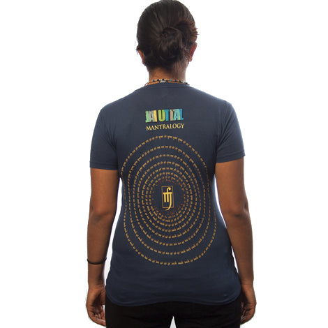 A person with dark hair in a ponytail wears a navy blue organic cotton Mantralogy/Gaura Vani Sadbhuj shirt with MUTAL MANTRALOGY and a circular golden design with text on the back, standing against a plain white background.