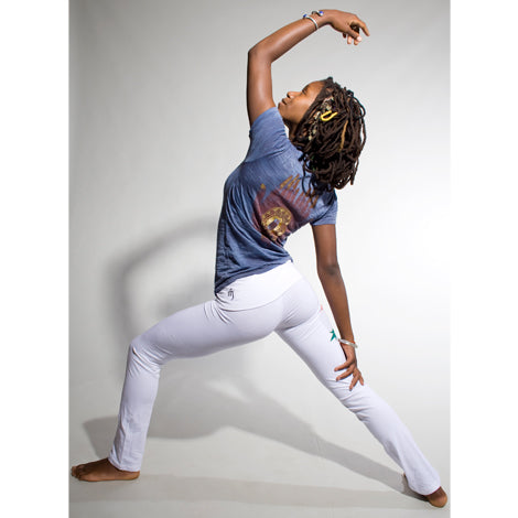 A woman with dreadlocks performs a yoga pose barefoot on a white surface, wearing a blue tee and Mantralogy summer-weight cotton yoga pants with a fold-over waistband, one arm reaching overhead and the other on her thigh.