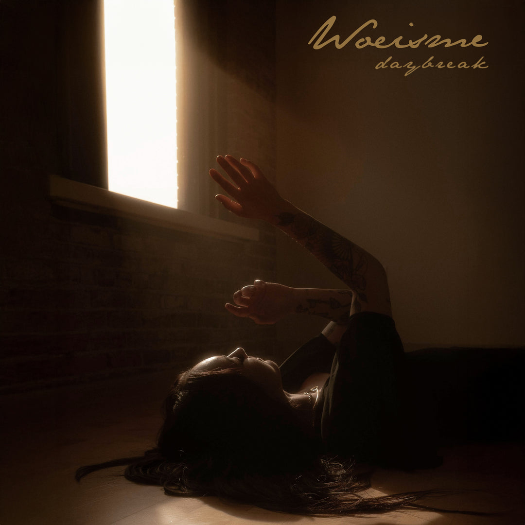 A person lies on a wooden floor in a dimly lit room, reaching toward sunlight through a window. Woe Is Me Daybreak Album Presale is displayed in the upper right corner.