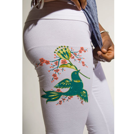 A person wearing Mantralogy white yoga pants with a fold-over waistband and a colorful green hummingbird and flower design on the upper thigh, with their torso and hand partially visible.