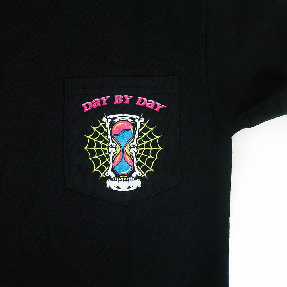 close up of the pocket of the shirt has a hourglass with a spiderweb behind it and the text Day By Day in pink above the hourglass.