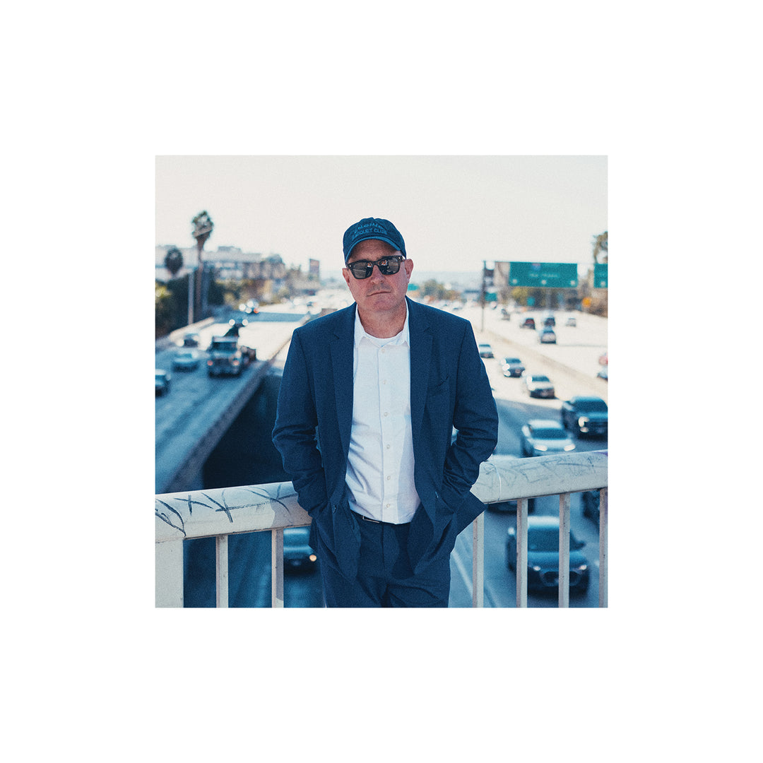A man in a blue suit, white shirt, sunglasses, and dark baseball cap stands on a bridge above a busy freeway in daylight—channeling the cool vibe of Craig Finn.