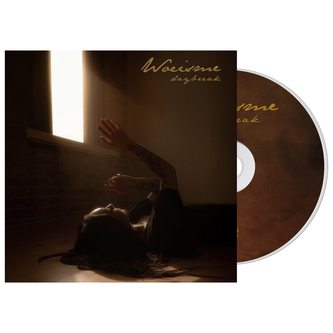 A Woe Is Me CD case features someone on a wooden floor, reaching for sunlight through a window. The Daybreak album title appears in gold script at the top—ideal for fans looking for a presale music bundle.