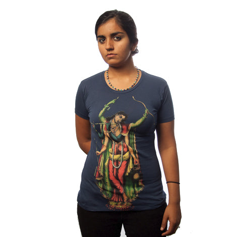A woman with a serious expression stands against a white background, wearing a blue organic cotton Sadbhuj shirt from Mantralogy/Gaura Vani. The shirt features a vibrant artistic image of Sri Chaitanya playing a flute. Her hands are clasped behind her back.