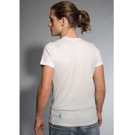 A person with long brown hair in a ponytail faces away from the camera, wearing a Mantralogy short-sleeved off-white organic cotton T-shirt and blue jeans, set against a plain gray background.
