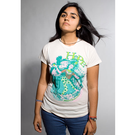 A person with long dark hair faces forward, wearing a Mantralogy limited edition organic cotton light tee featuring a colorful HOPE graphic, paired with blue jeans and a beaded necklace, set against a plain background.
