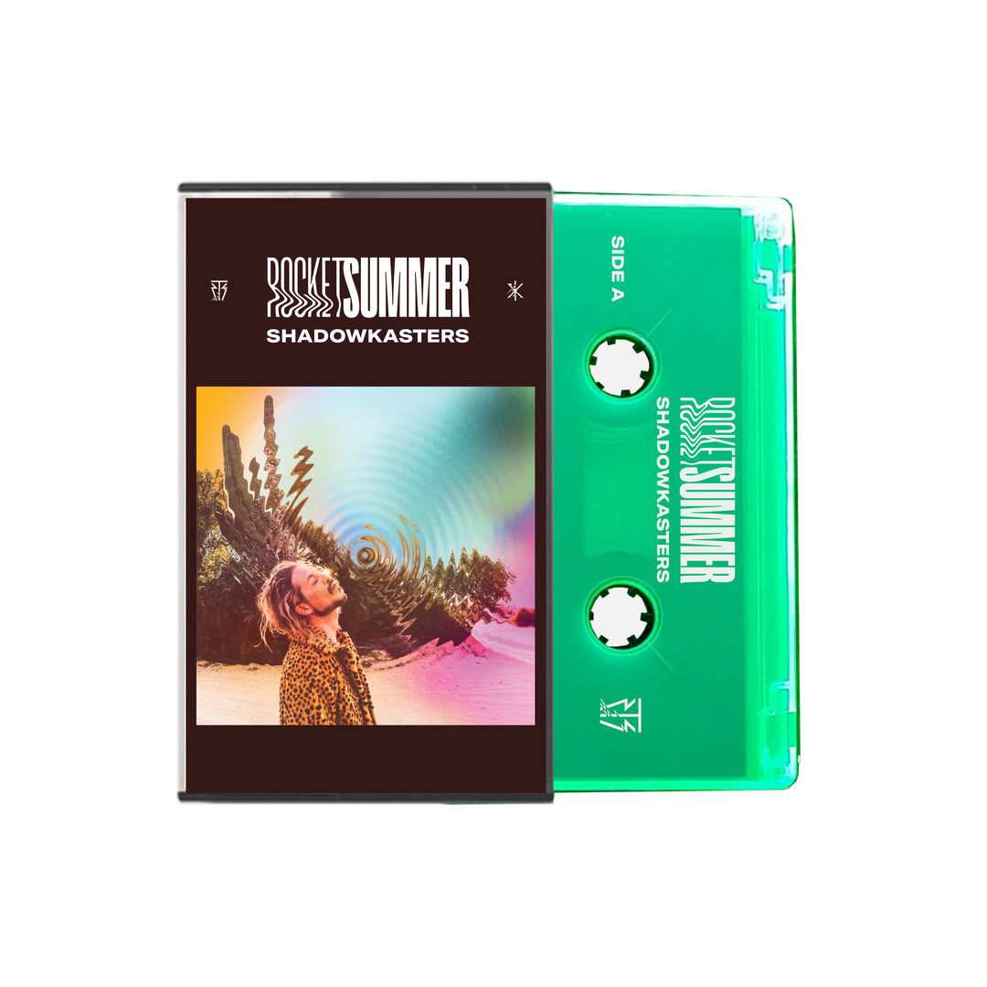 A glow-in-the-dark, fluorescent green cassette tape from The Rocket Summer sits beside its case, which displays vibrant artwork of a sunglasses-wearing figure in a sunlit desert with cacti and radiating rainbow patterns.