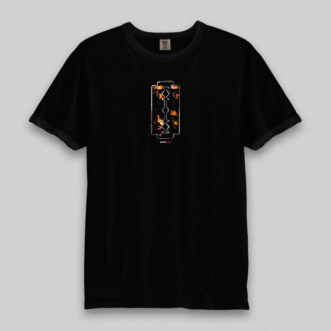 Boundaries’ black garment-dyed tee showcases a vertical razor blade graphic filled with orange and yellow flames. The relaxed fit shirt is presented on a light, neutral background.