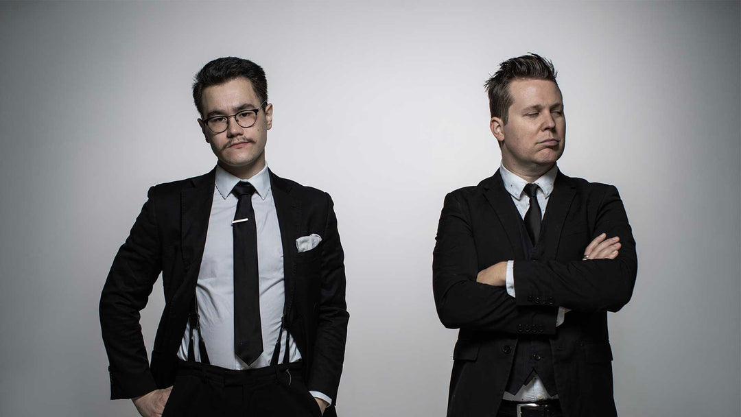 Two men in suits stand against a plain gray background; one faces the camera with hands on hips, while the other stands with arms crossed, eyes closed, looking to the side.