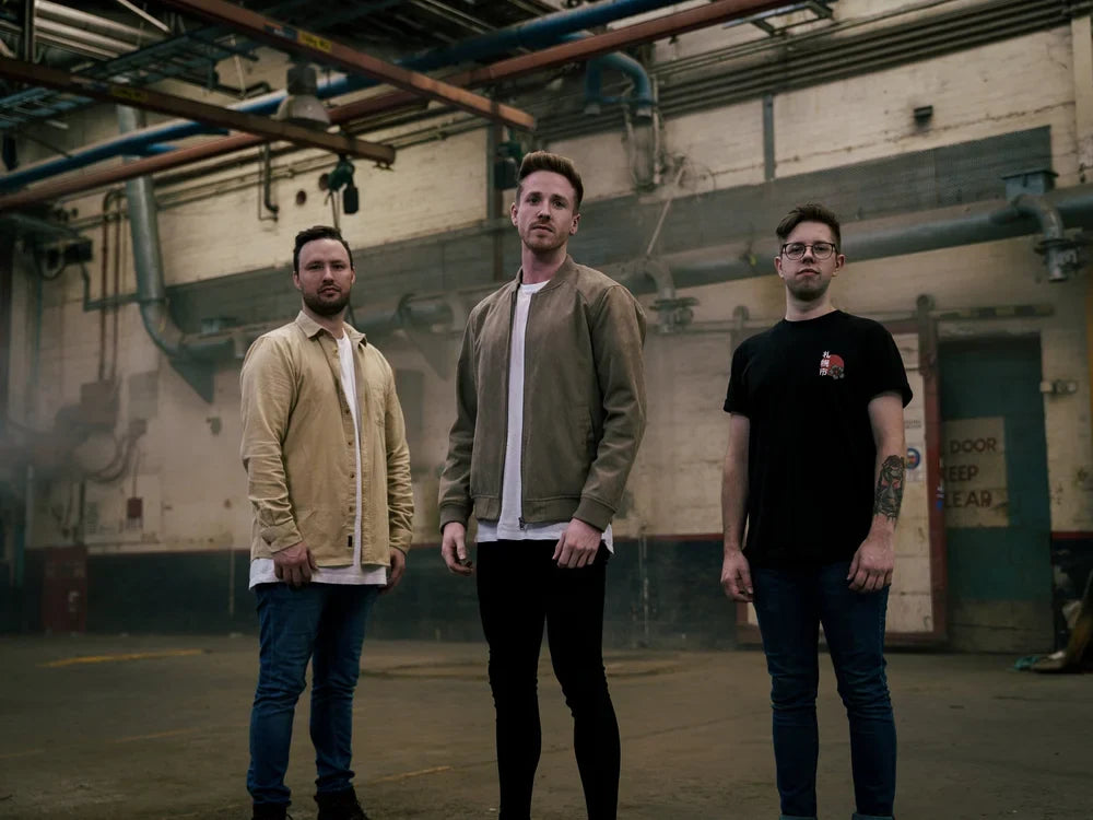 Three men stand in an industrial warehouse, facing the camera. Two wear light-colored jackets and one wears a black T-shirt. Exposed pipes and worn walls are visible in the background.