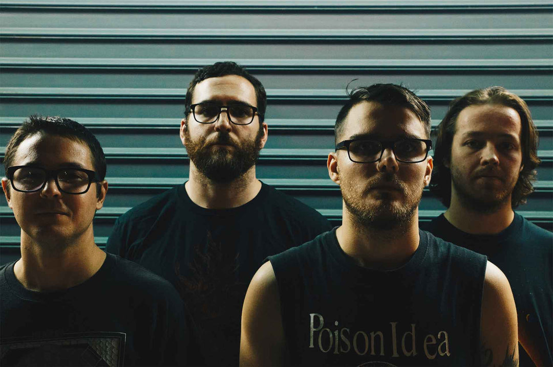 Four men with serious expressions stand in front of a metal shutter, wearing dark clothing and glasses, with dim lighting casting strong shadows on their faces.