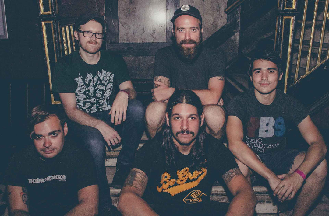 Five men sit on marble stairs indoors, facing the camera and smiling. They wear casual t-shirts, and one has a beard and cap. The setting has dark walls and gold accents.