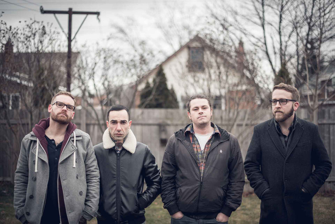 Four men stand side by side outdoors in a yard with bare trees and houses in the background, all wearing coats and looking toward the camera with neutral expressions.