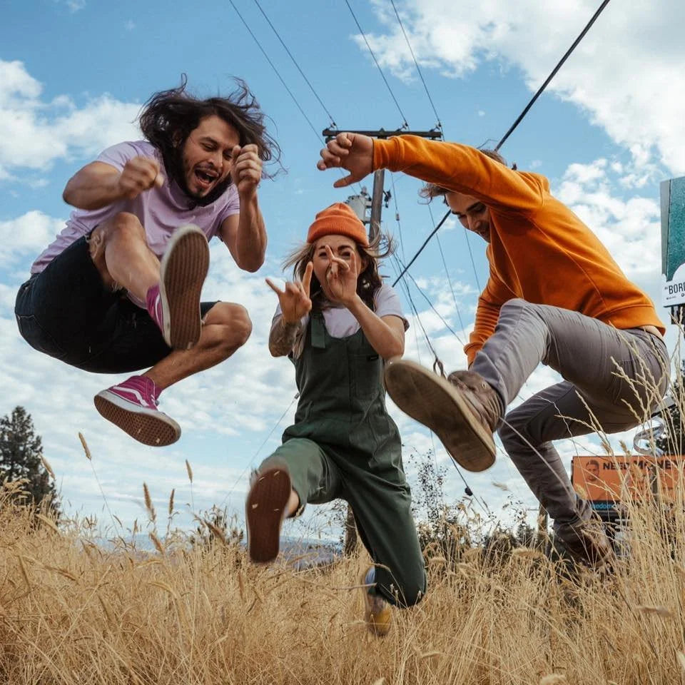 Three people jump enthusiastically in a field of tall grass under a blue sky with clouds, captured mid-air with joyful expressions and dynamic poses.