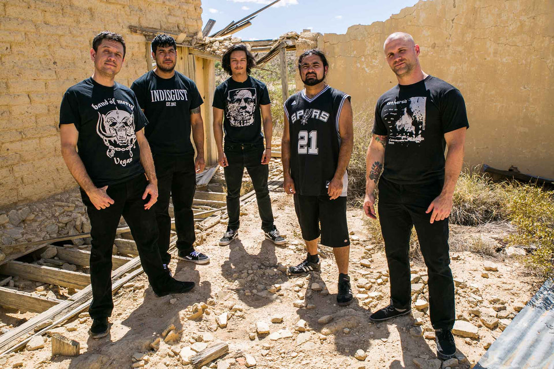 Five men wearing dark clothing stand in a sunlit, abandoned, partially collapsed building with rubble and wooden debris on the ground around them.