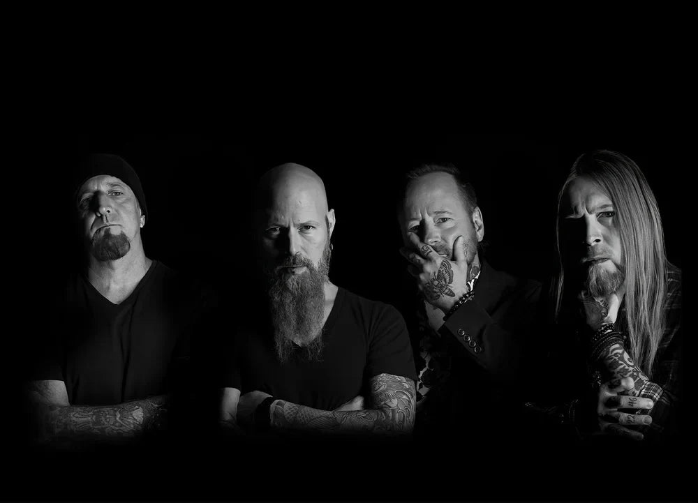 Four men with tattoos pose in a black-and-white photo against a black background. Three have beards, and one is clean-shaven with long hair. All appear serious or contemplative.
