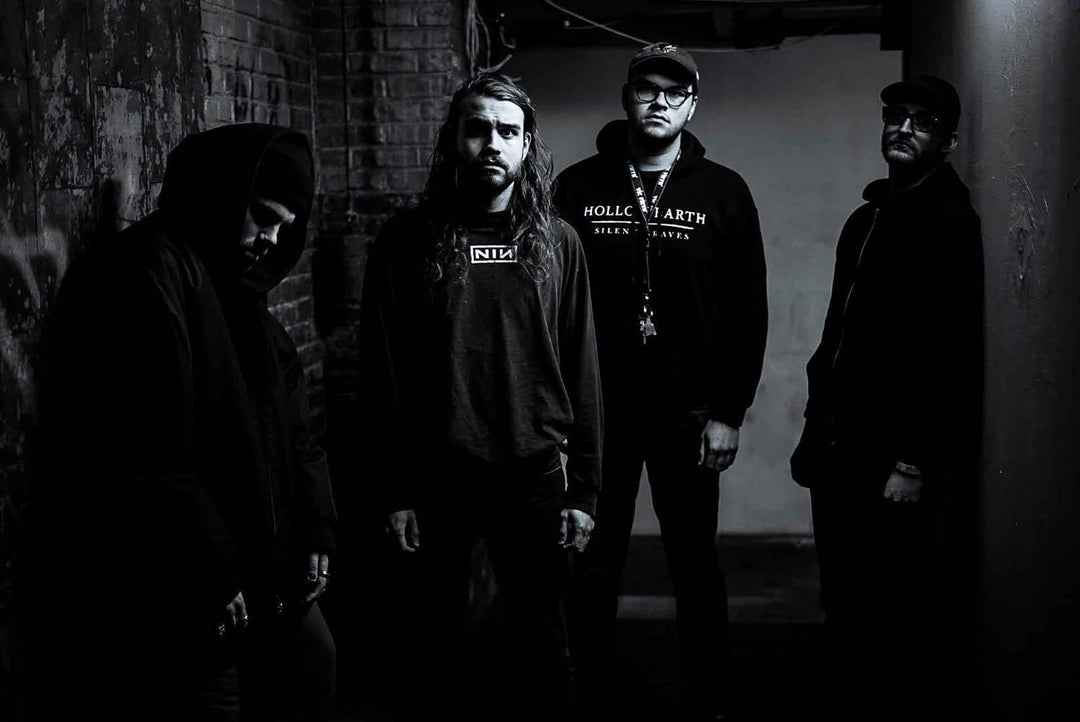 Four men in dark hoodies and jackets stand in a dimly lit, narrow alley with brick walls, looking serious and facing the camera. The image is in black and white.