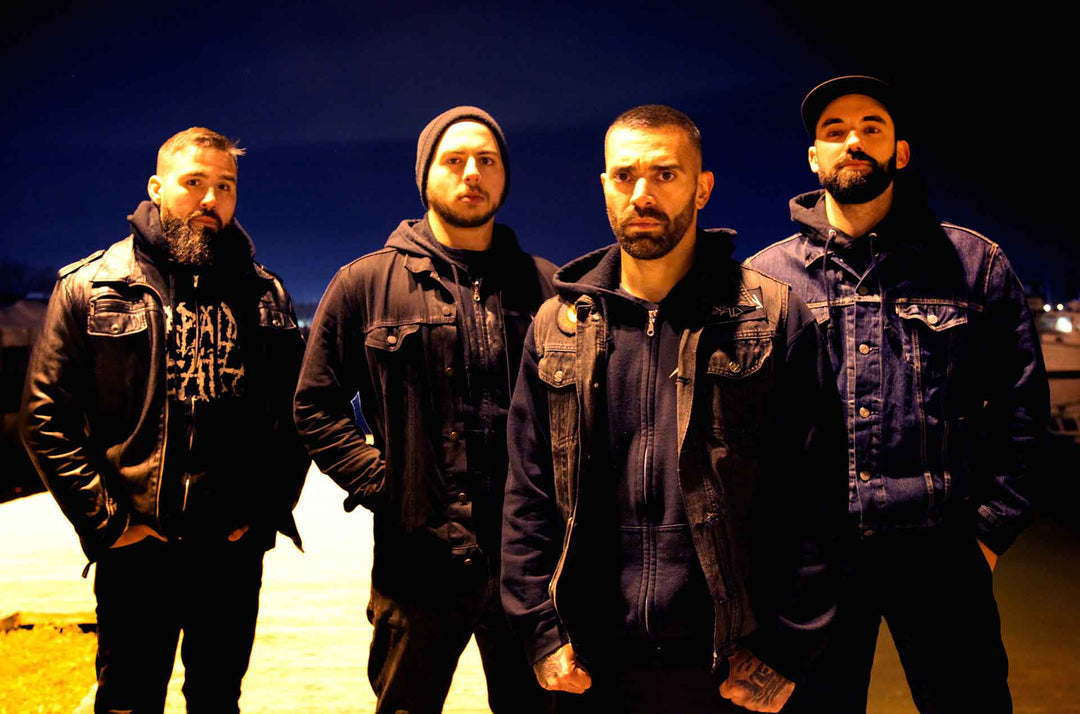 Four men with beards, wearing dark jackets and standing outdoors at night, look seriously at the camera under artificial lighting.
