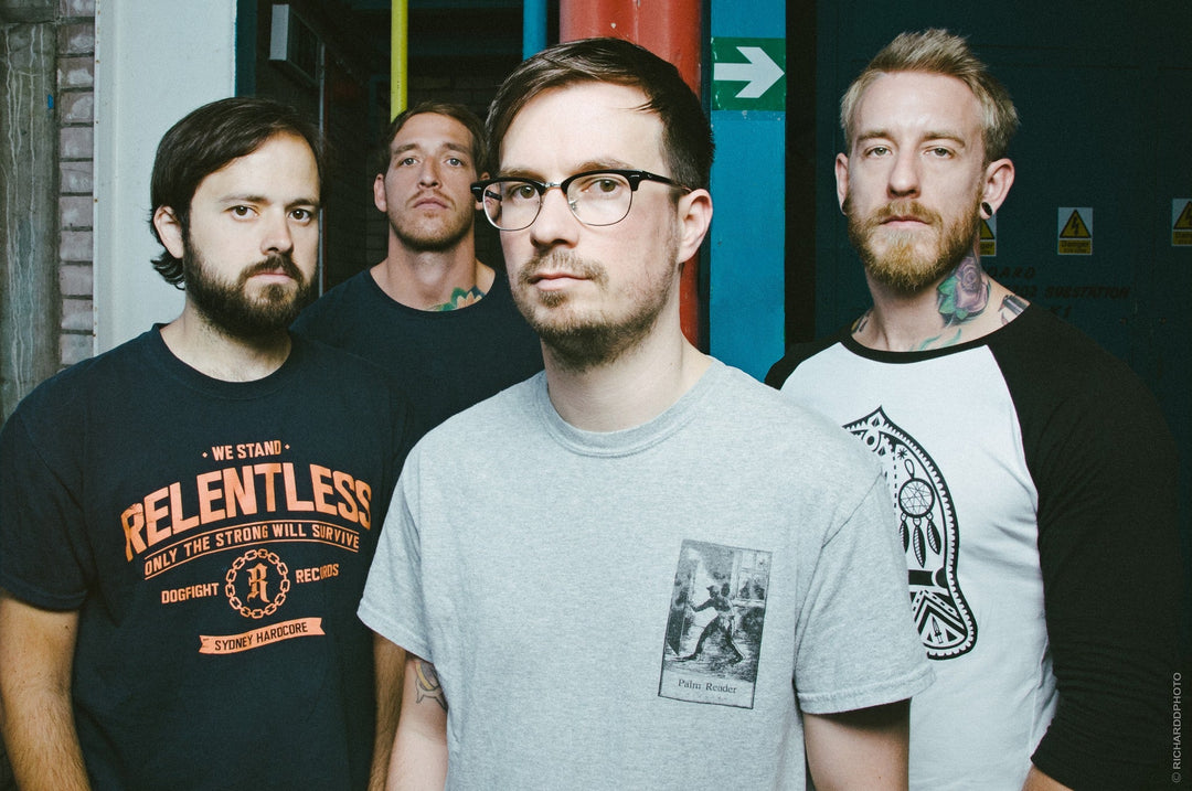 Four men stand together, three with beards and one with glasses, with Funeral for a Friend written in script across the image. The background includes a green exit sign and industrial elements.