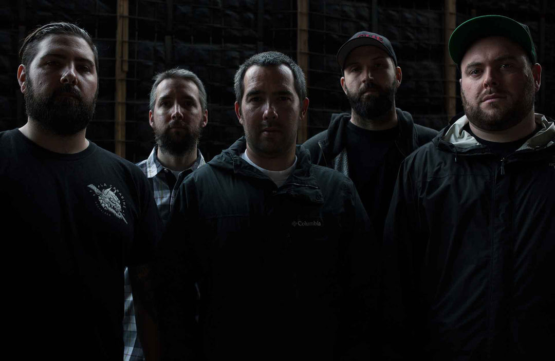Five men with beards and serious expressions stand closely together in dim lighting, wearing dark casual jackets and hats, with a dark background of metal bars and wooden beams.