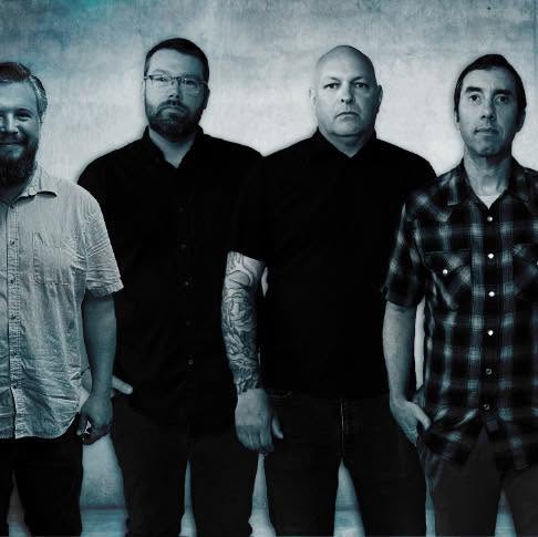 Four men stand side by side against a blue-gray backdrop. Three wear dark shirts, and one wears a light short-sleeve shirt. One man has a visible arm tattoo.