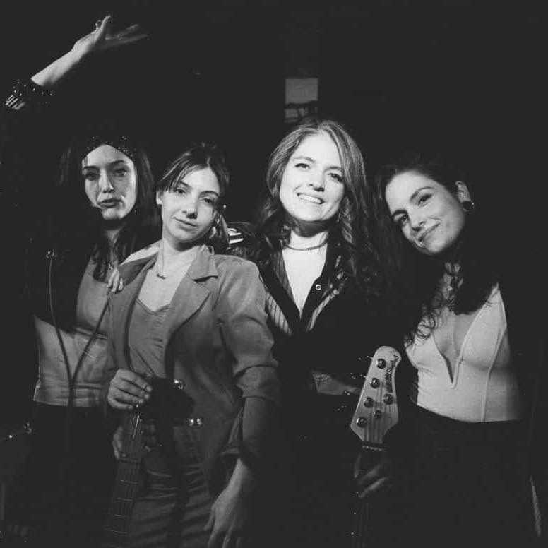 Four women stand together, smiling and posing with electric guitars, wearing stylish outfits in a black-and-white photo with a dark background.