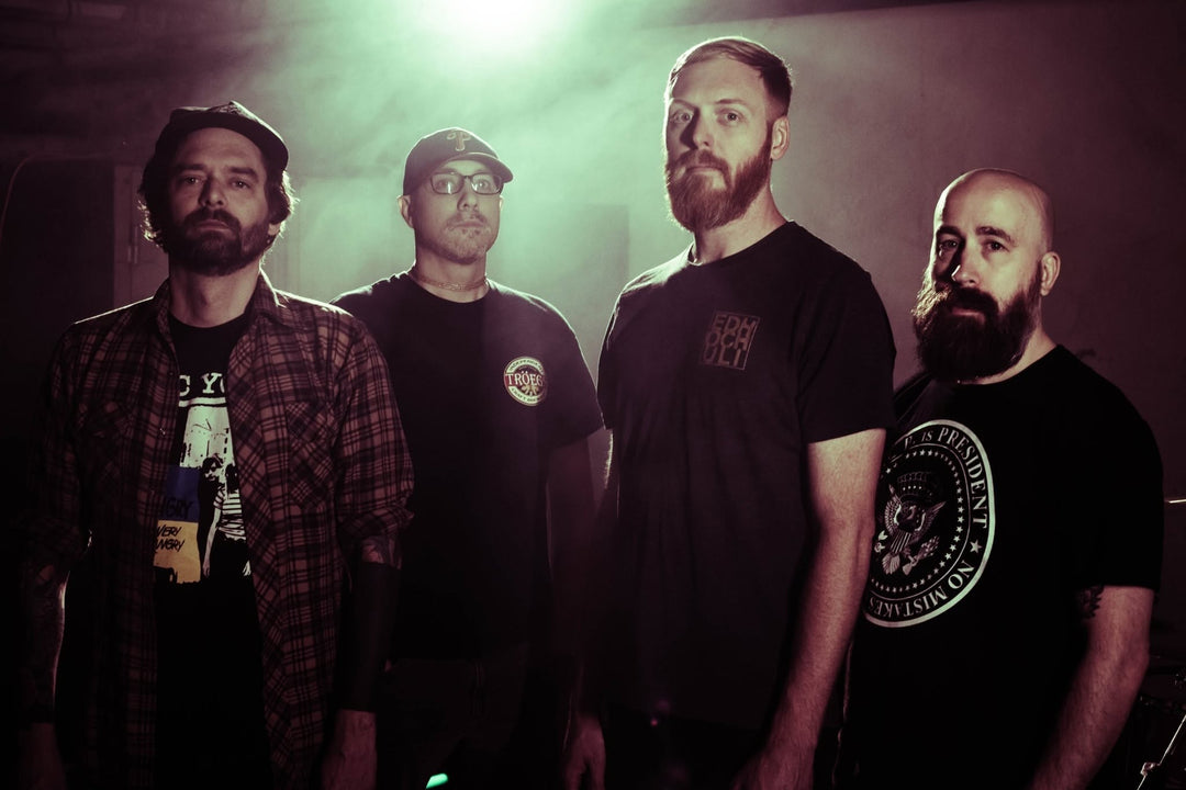 Four men stand together in dim lighting, all with serious expressions. They wear casual clothes, including t-shirts and hats, and have beards. A bright light shines behind them.