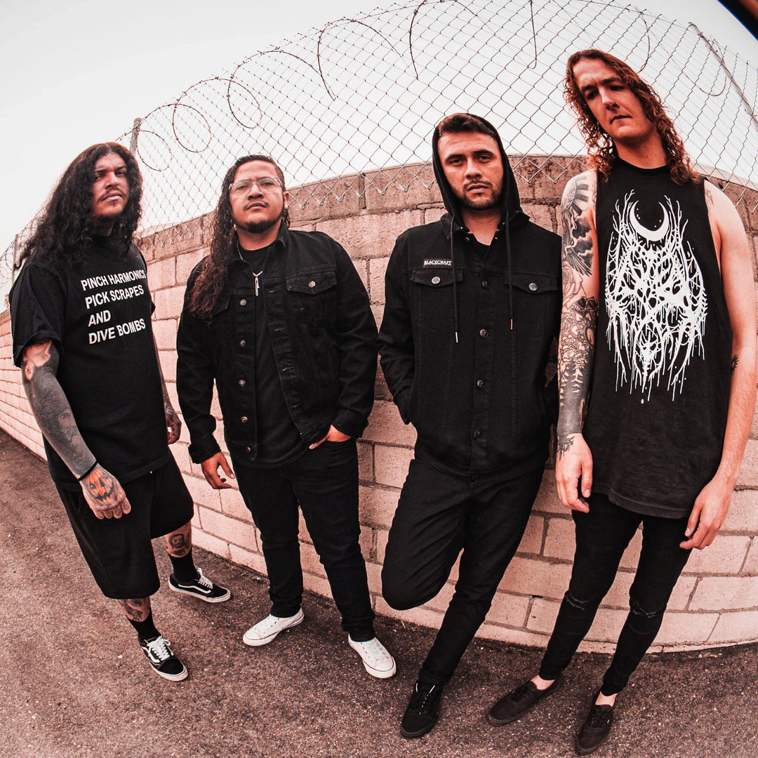 Four men with long hair and tattoos stand in front of a brick wall topped with barbed wire, dressed mostly in black casual and band clothing, looking seriously at the camera.