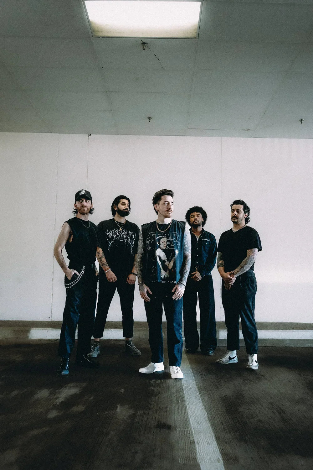 Five men with tattoos stand in a row in an urban setting, wearing dark casual clothing and looking confidently at the camera. Bright, natural light highlights the group.
