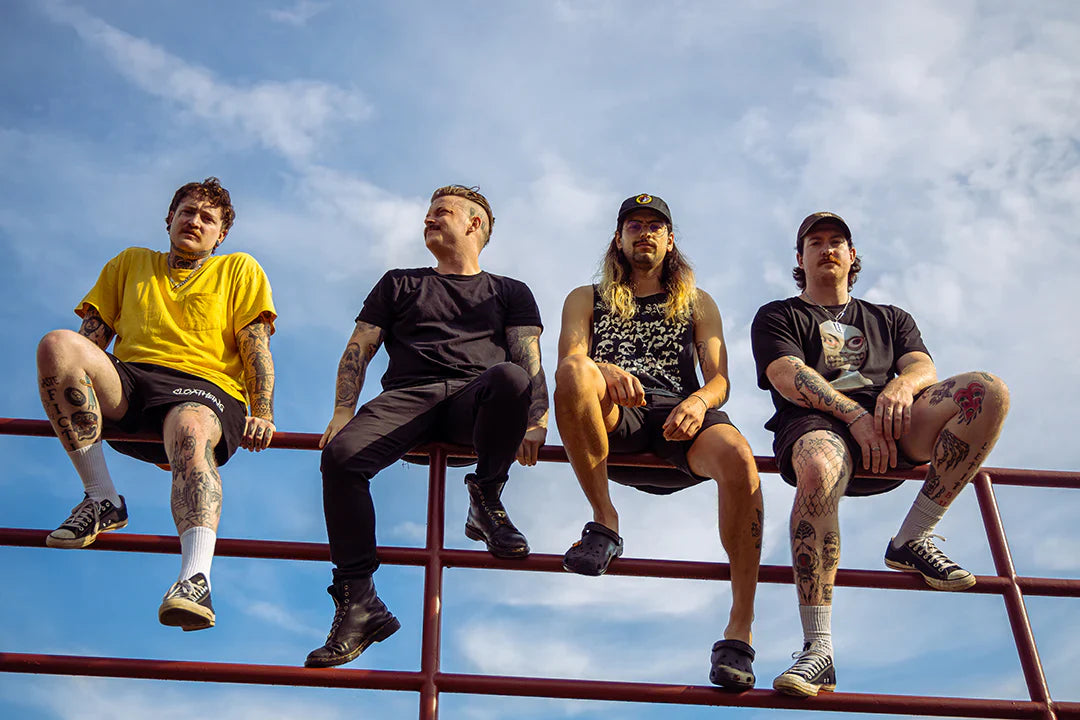 Four men with tattoos sit on a red metal railing against a blue sky, wearing casual clothes including t-shirts, shorts, and sneakers. Two wear hats, and all appear relaxed and look in different directions.