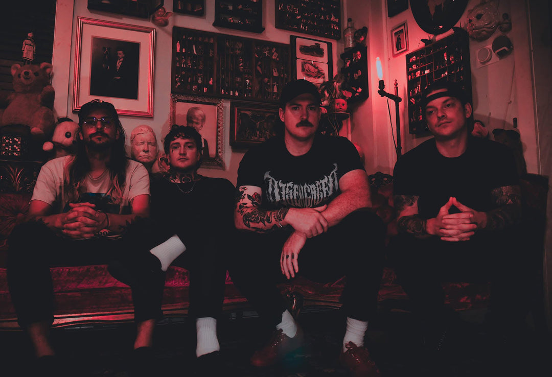 Four men with tattoos sit on a red metal railing against a blue sky, wearing casual clothes including t-shirts, shorts, and sneakers. Two wear hats, and all appear relaxed and look in different directions.