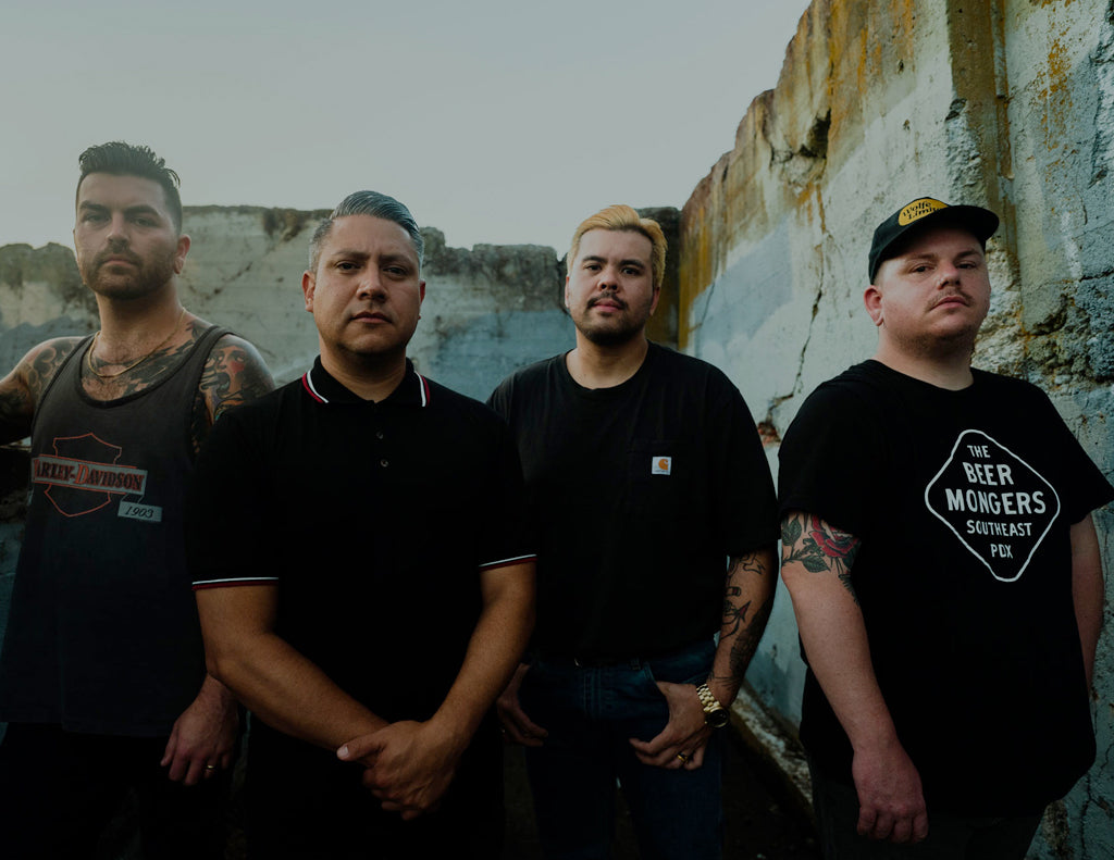 Five men pose for a photo next to and inside a gray van. They wear casual clothing, including graphic T-shirts, shorts, and caps. Some are seated, and some are standing, looking at the camera and smiling gently.