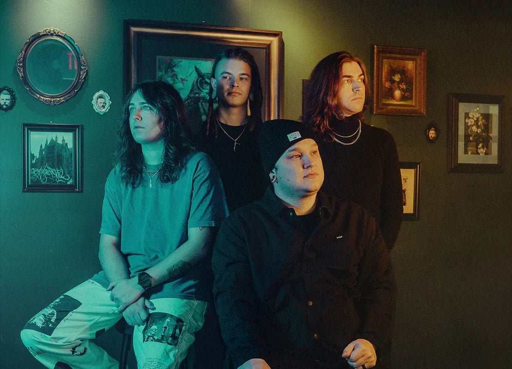 Four people with long hair pose indoors against a wall decorated with framed art and portraits. Three stand in back, one sits in front. The lighting is blue and yellow, creating a moody atmosphere.