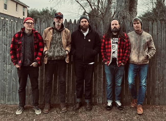 Five men stand side by side in front of a wooden fence and trees, dressed in casual jackets and jeans, some wearing hats, with neutral expressions.