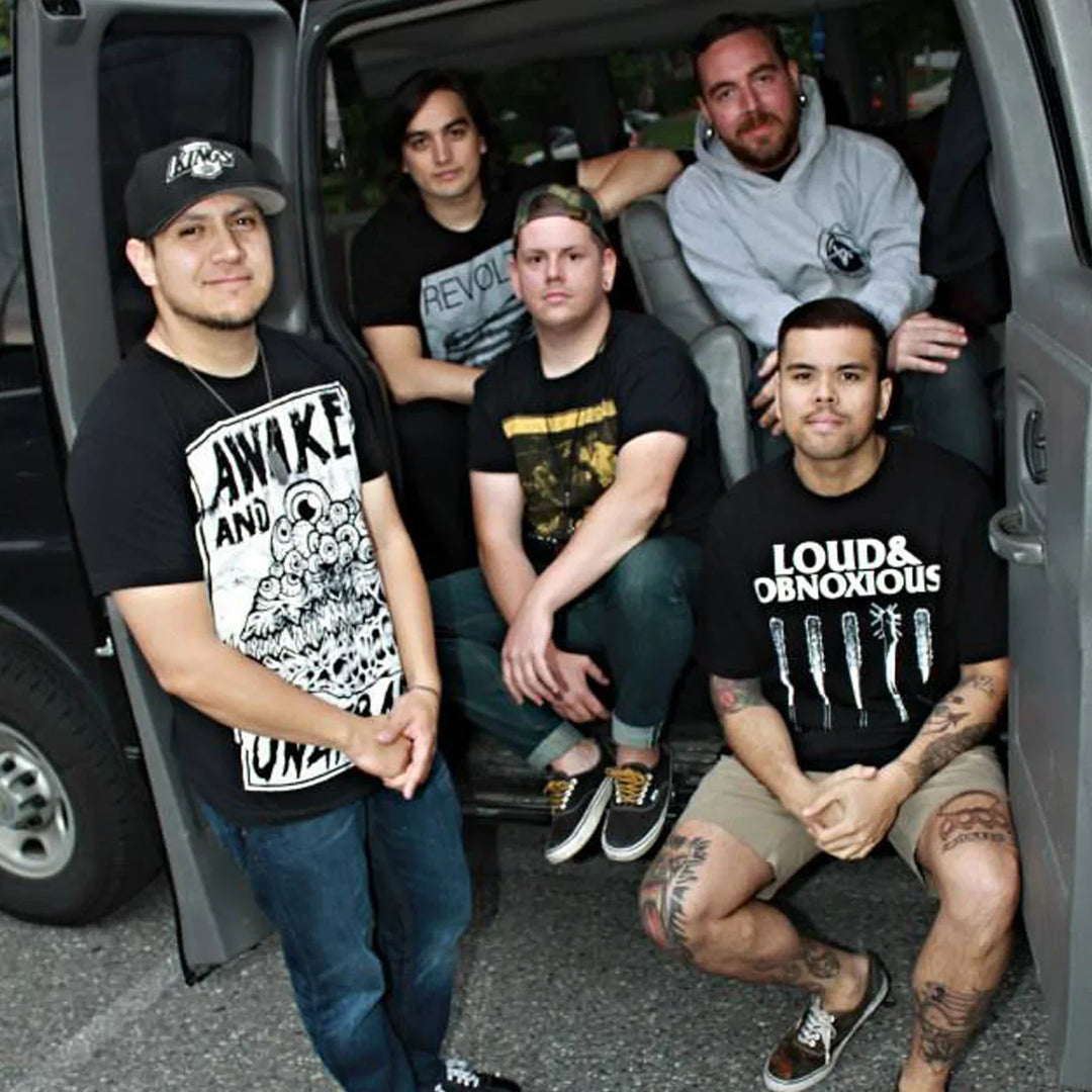 Five men pose for a photo next to and inside a gray van. They wear casual clothing, including graphic T-shirts, shorts, and caps. Some are seated, and some are standing, looking at the camera and smiling gently.
