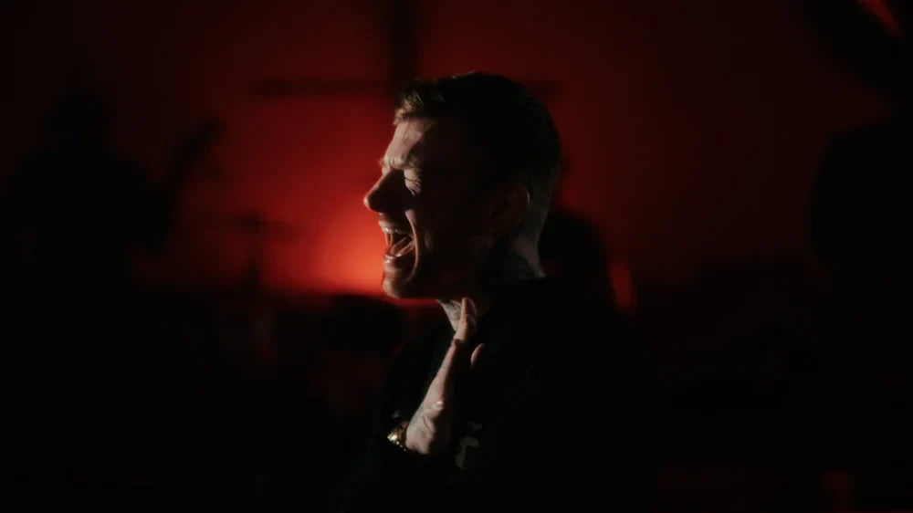 A person with short hair passionately singing or shouting, illuminated by dramatic red and black lighting with shadows in the background.