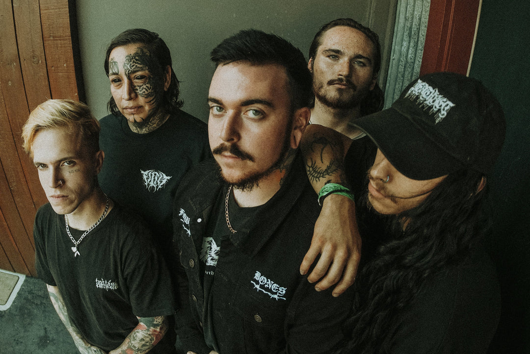 Five people with serious expressions pose closely together, wearing black clothing with band logos. One person has facial tattoos, another wears a black cap, and all display tattoos on their arms or necks.