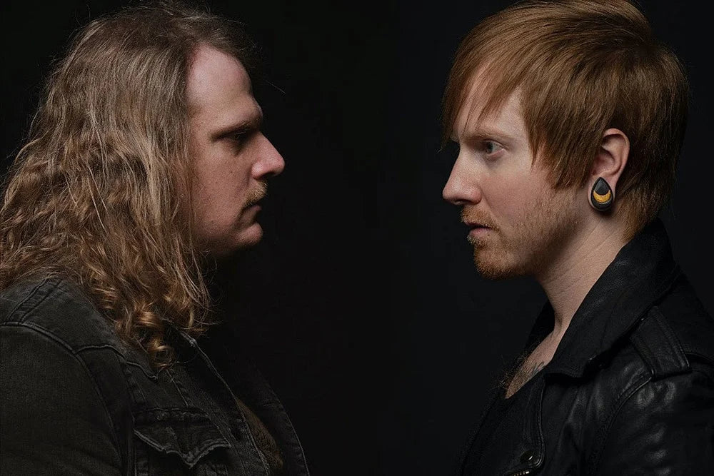 Two men face each other against a black background. One has long curly hair and a mustache; the other has short red hair, a beard, and large black ear gauges. Both wear dark jackets.