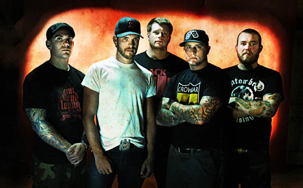 Five men with serious expressions stand side by side in front of a red-orange background, wearing casual clothing and caps, with visible tattoos on their arms.