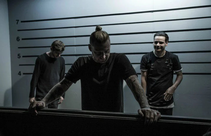 Three men stand in front of a height measurement wall, resembling a police lineup. One man leans on a railing, while the others stand behind him, all dressed in dark clothing under dim lighting.