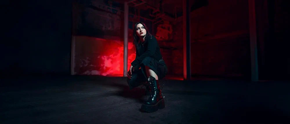 A woman dressed in black crouches confidently in a dark, industrial space lit with dramatic red light in the background. She wears chunky black boots and gazes forward.
