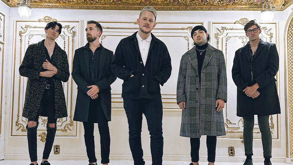 Five men stand in a row in a lavish, ornate room with white walls and gold trim, all wearing stylish coats and dark clothing, looking confidently at the camera.