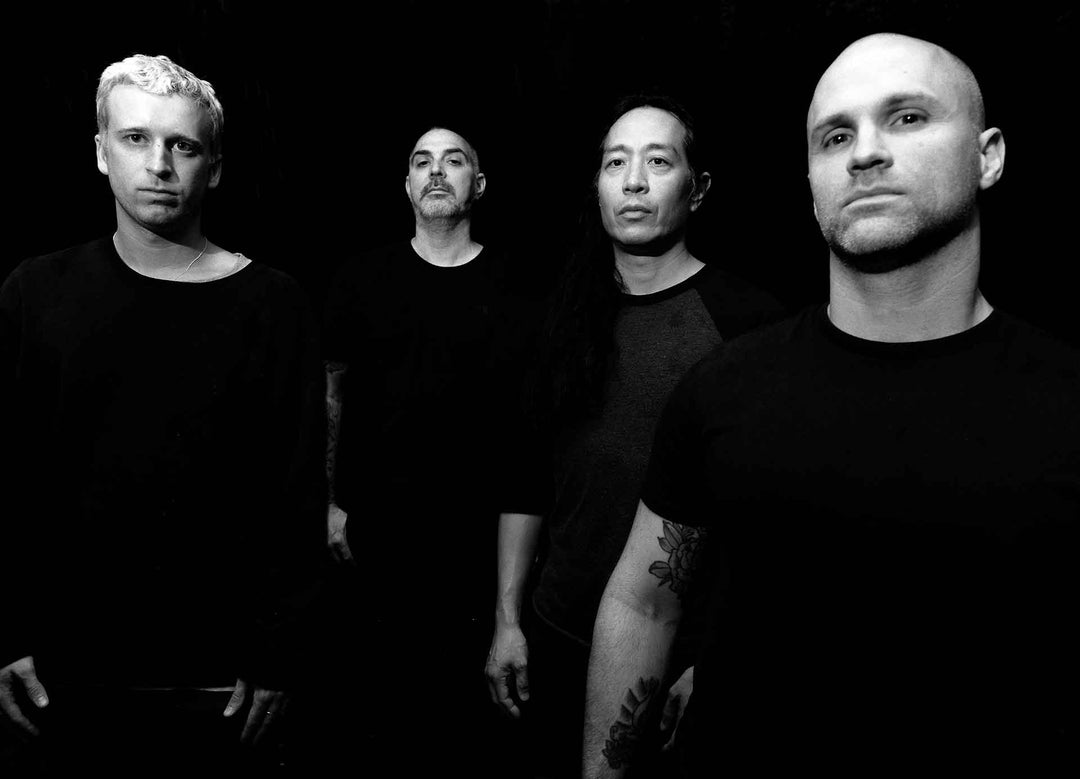 Four men stand side by side in a black and white portrait against a dark background, all wearing dark shirts and looking toward the camera with serious expressions.
