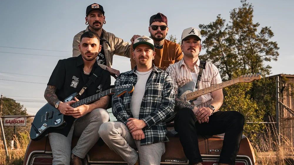 Five men pose outdoors on the back of a vintage car, holding electric guitars. They wear casual clothes and hats, with trees and a building in the background under a clear sky.