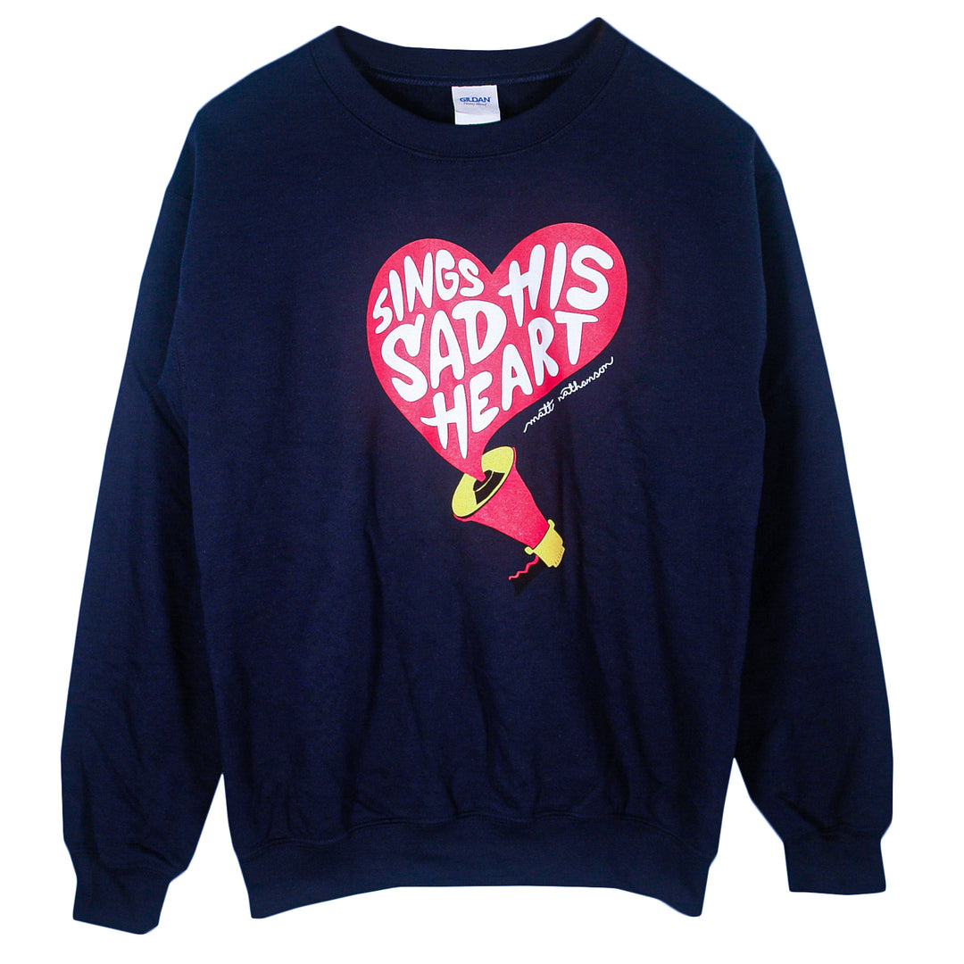 A navy blue Matt Nathanson Gildan Heavy Blend crewneck sweatshirt features a red heart with the white text "Sings Sad His Heart" inside, plus a yellow and red megaphone below, and a small signature beneath the graphic.