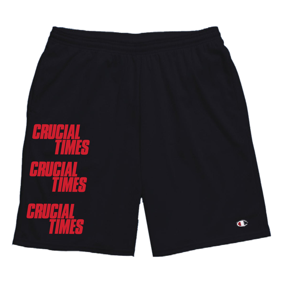 Black mesh shorts from Crucial Times featuring "CRUCIAL TIMES" printed three times in bold red down the left leg. A small Champion logo appears near the right hem. Available now for presale.