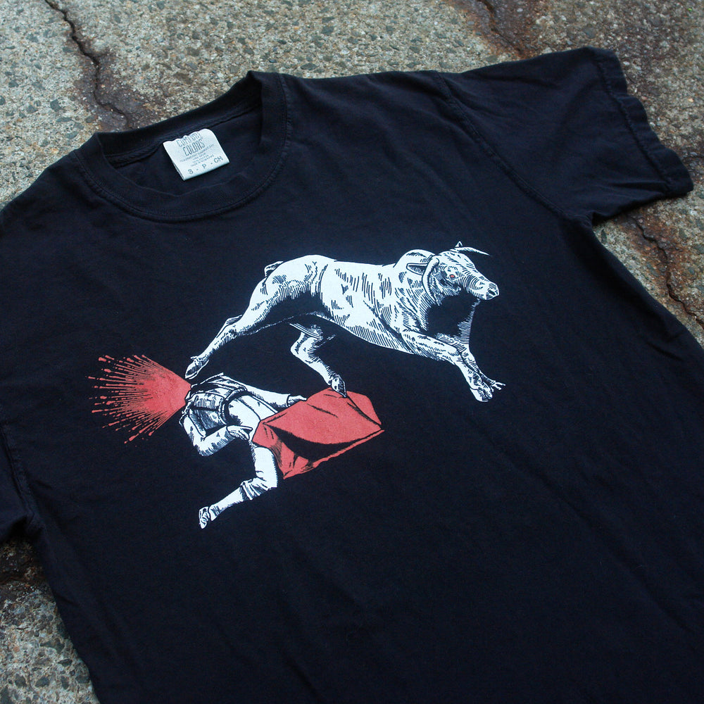 The MerchNow black garment-dyed T-shirt displays a bull goring a matador, who falls forward with a burst of red from his head. The matador’s red cape stands out against the tee, shown laid on a rough surface.