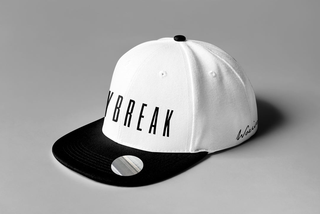 A Woe Is Me baseball cap with a white crown, black brim and button, BREAK in bold black letters on the front, and a subtle Daybreak merch reference in cursive on the side, shown on a plain gray surface.