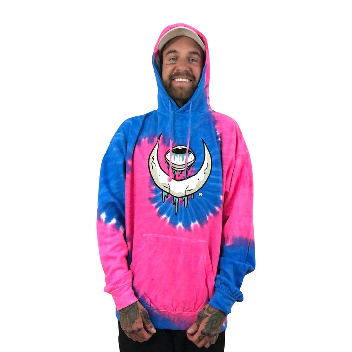 A smiling person wears a Stay Sick Clothing pink and blue tie-dye hoodie featuring a large crescent Eye Moon and character graphic on the front, standing against a plain white background.
